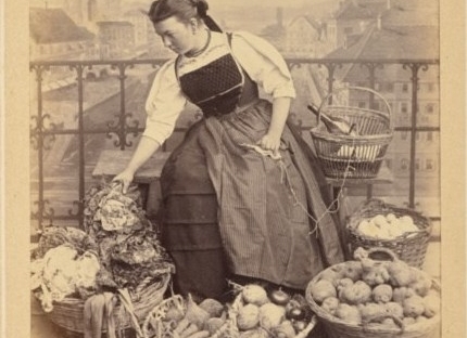 19th century photo of Swiss woman knitting, surrounded by baskets of vegetables.