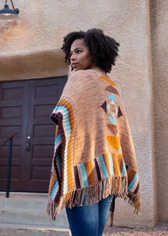 Woman wearing poncho style cardigan with geometric intarsia colour-work details and fringe edging, in warm colours inspired by the American Southwest.