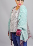 Woman wearing oversized, slouchy shrug, knitted in merino/mohair In multicoloured, striped brioche sections, with contrast colour edging and cuffs.