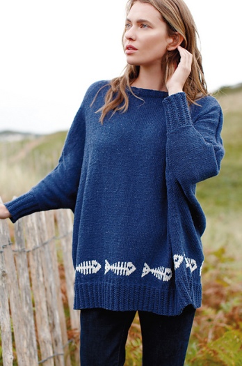 Woman wearing oversized, knitted sweater in dark blue with repeating motif of fish skeletons in white that run above the ribbed hem.
