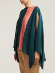 Woman wearing knitted cape with open sleeves and boat neck in teal shade, with contrasting stripe of peach running vertically across one side of garment.