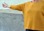 Woman, with arm outstretched, wearing mustard yellow, knitted sweater with all-over bobble texture.