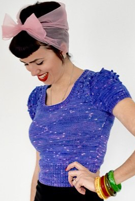 Woman wearing fitted, knitted top with bobble detail at shoulders and deep rib at hem. Styled with multicoloured bangles and tulle head scarf.