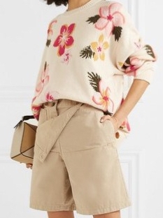 Model wearing oversized, cream sweater with multicoloured floral intarsia design; sweater worn tucked in to stone coloured shorts.