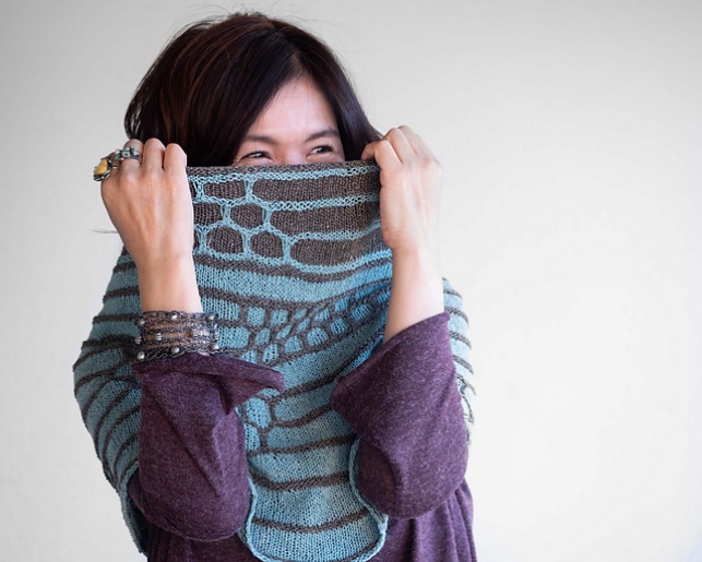 Woman wearing large, knitted cowl, pulled up over her face to show slip-stitch colour work design of curved sections of blue and grey colour.